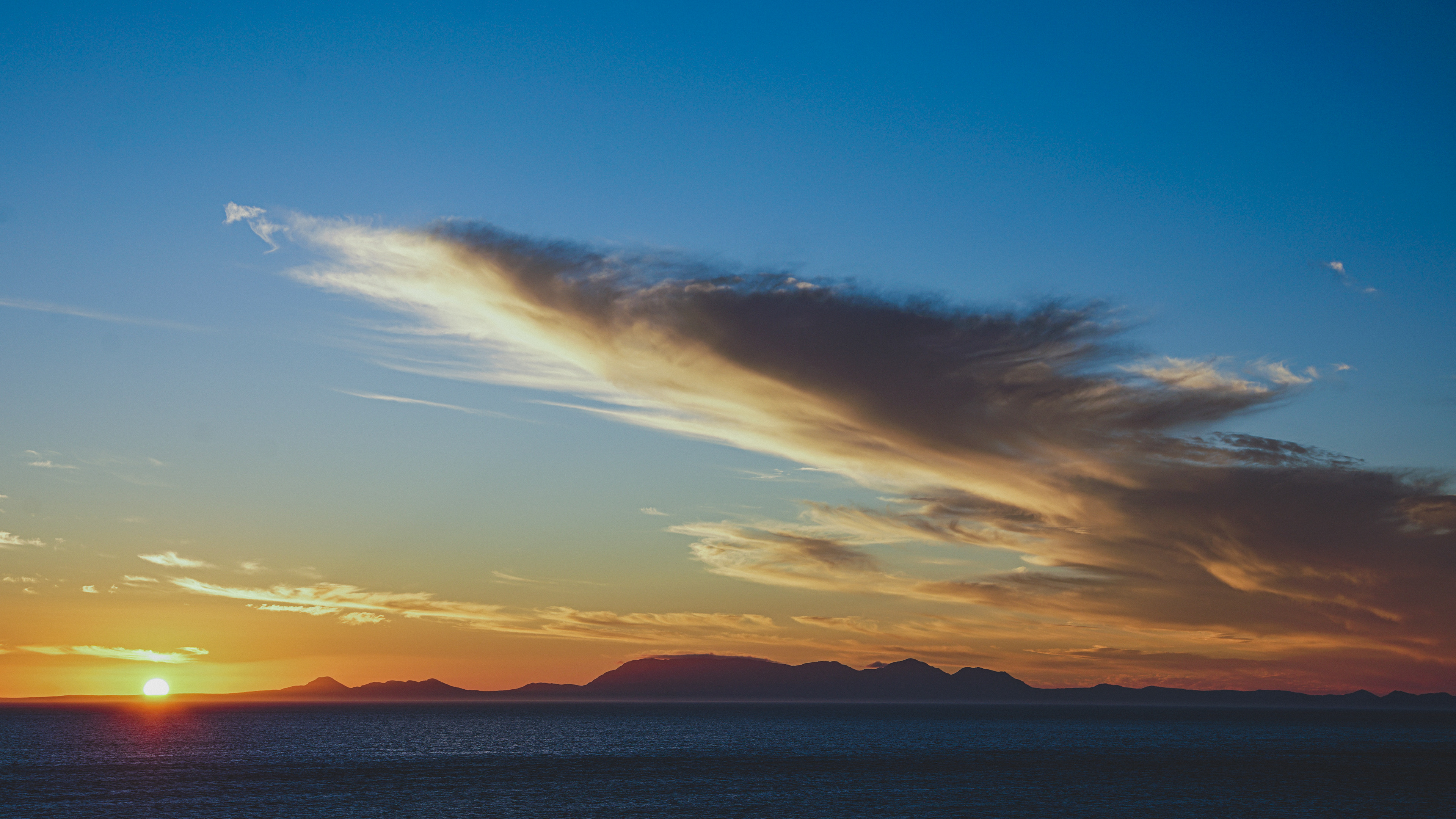 Vibrant sunset over ocean and silhouetted mountains, with dramatic orange-blue clouds in a gradient sky.